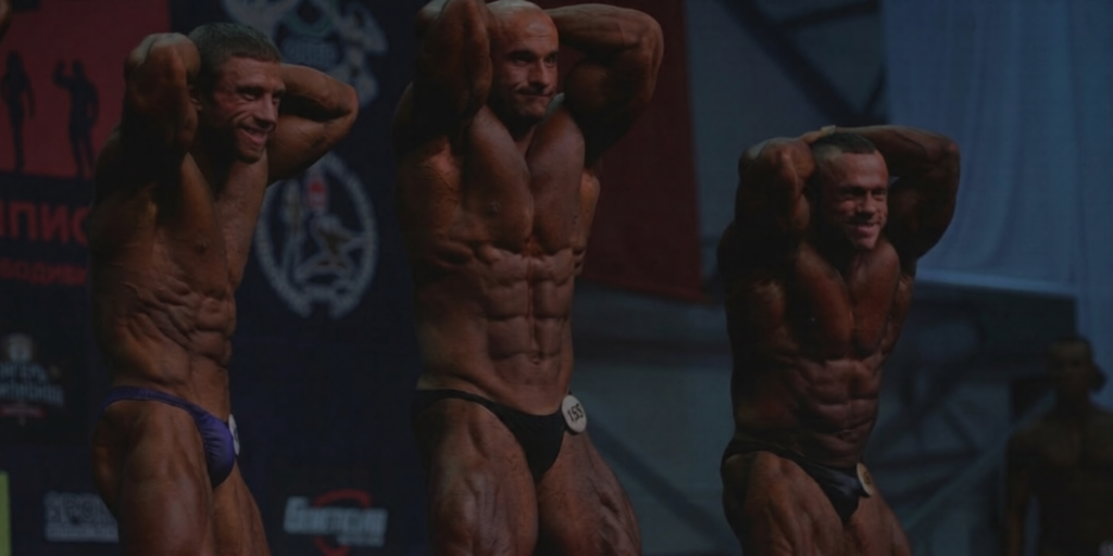 Tre manliga fitnessatleter poserar på scen under en bodybuilding-tävling. Scenfitness erbjuder tävlingslicens för atleter i hela Sverige.