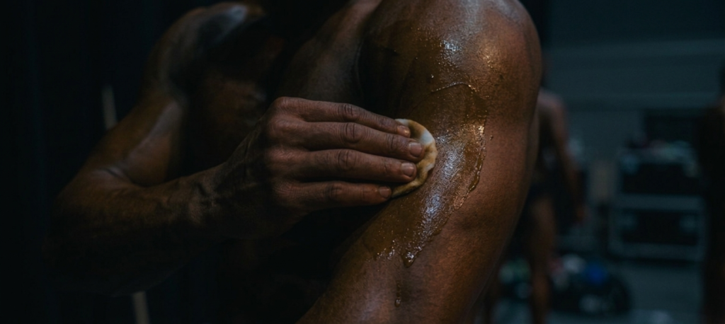 Här är en Google-optimerad alt-text för den nya, mörkare bilden som fokuserar på de sista förberedelserna:

alt="Närbild av en hand som applicerar tävlingsolja på en atlet backstage. Mörk och stämningsfull bild som fångar förberedelserna inför en fitness-tävling.